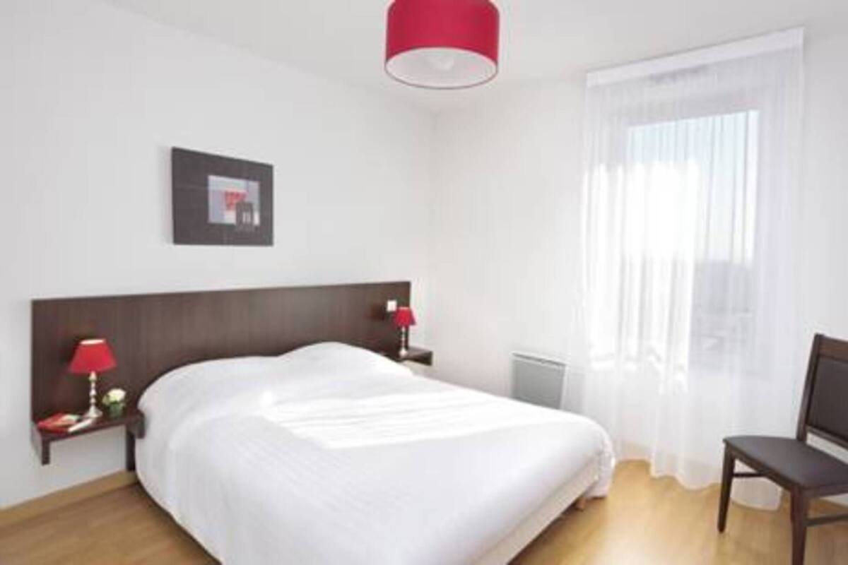 A hotel room with a bed, a nightstand with a lamp, a chair, a window with sheer curtains, and a picture on the wall.