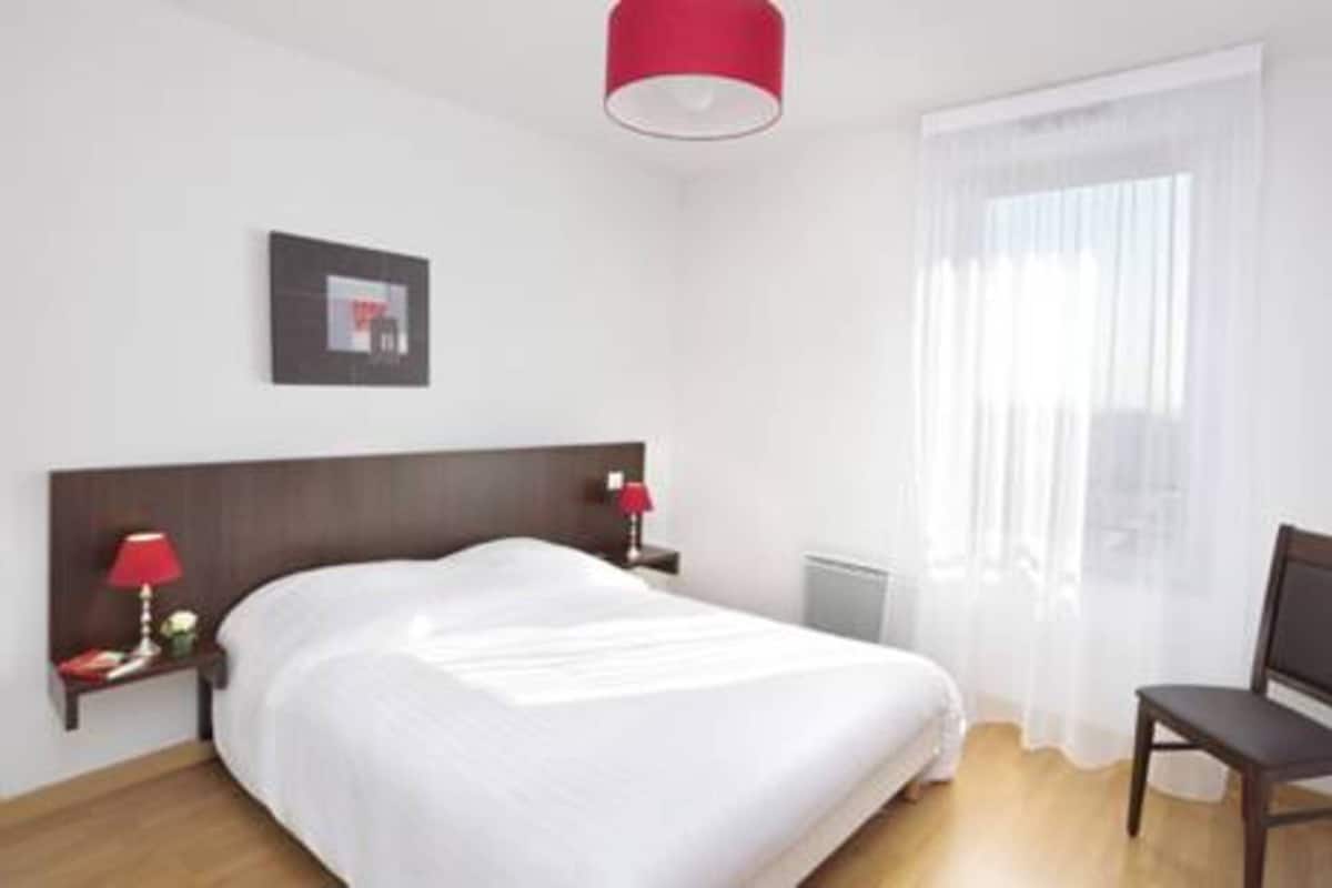 Une chambre d’hôtel avec un lit, une table de chevet avec une lampe, une chaise, une fenêtre avec des rideaux légers et un tableau au mur.