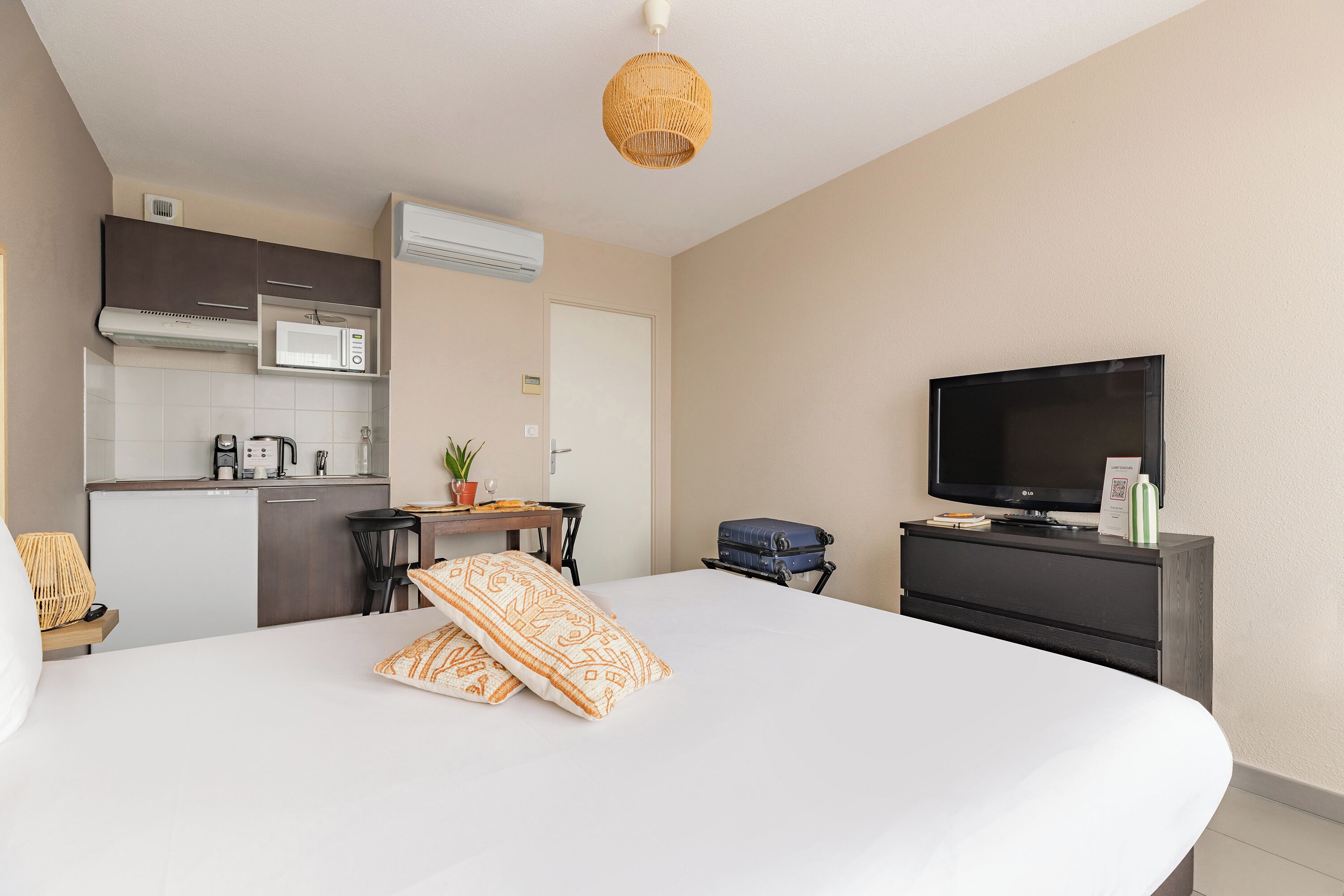 A hotel room with a bed, a TV, a desk with a microwave, a chair, and a suitcase.