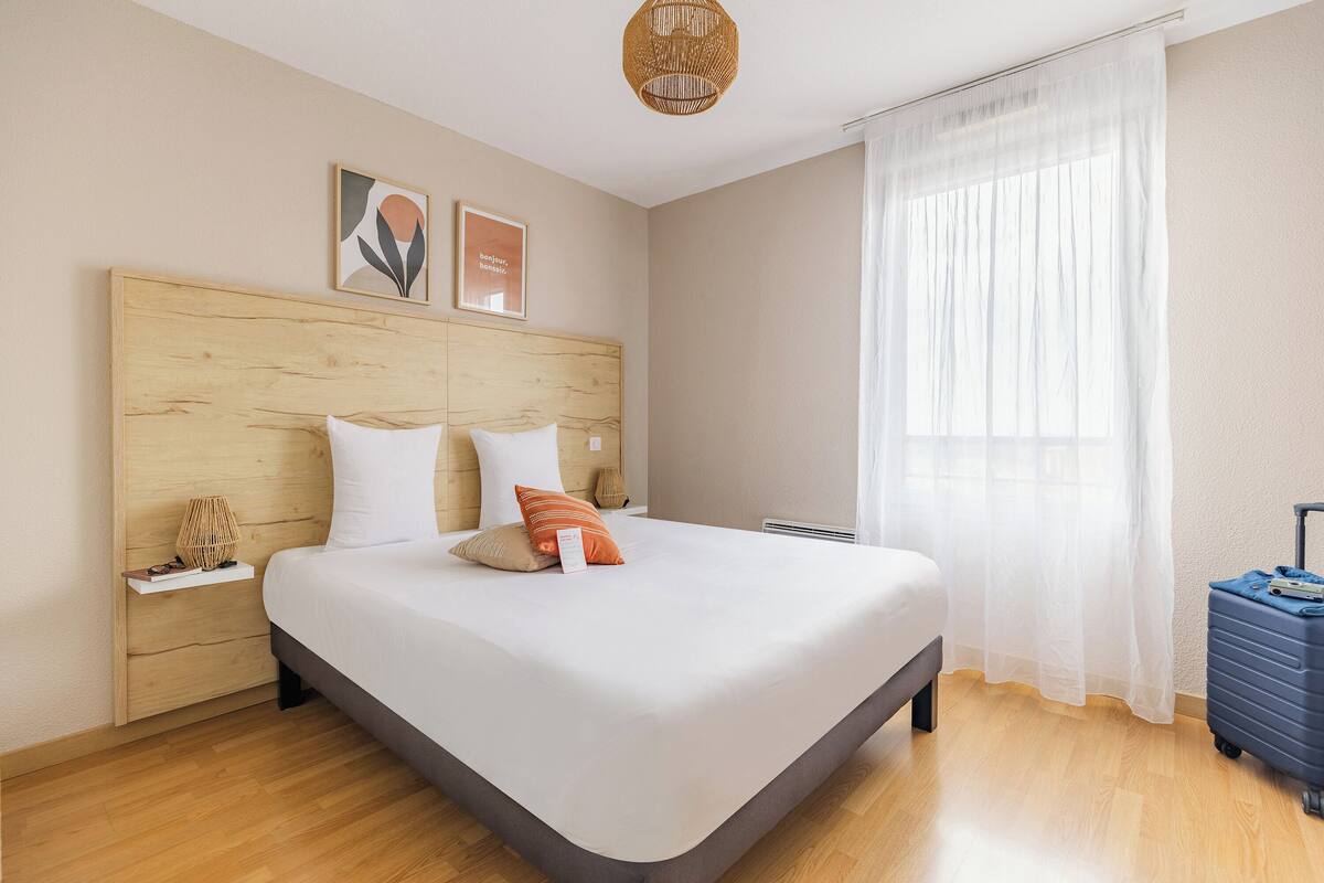A bedroom with a wooden headboard, a bed with white bedding, a nightstand with a lamp, a suitcase, and a window with sheer curtains.