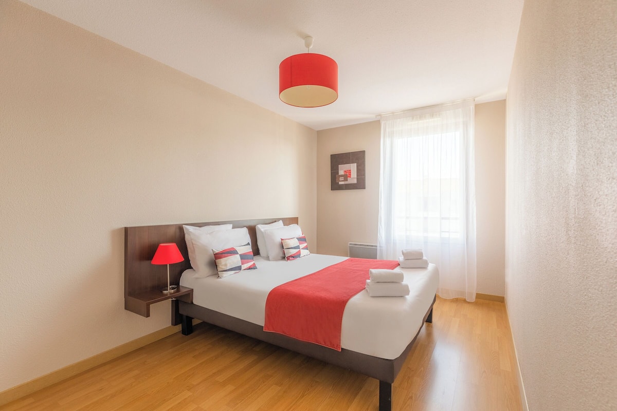 Une chambre d’hôtel moderne, équipée d’un lit, d’une table de chevet avec une lampe, d’une fenêtre avec des rideaux et d’un tableau accroché au mur.