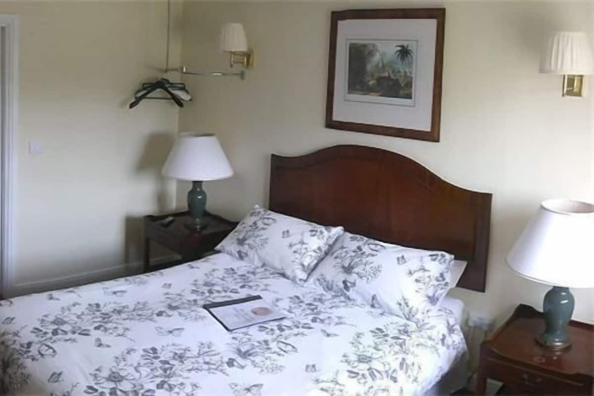 Quarto com cama, escrivaninha, cadeira, porta, televisão na parede, luminária e quadro emoldurado.