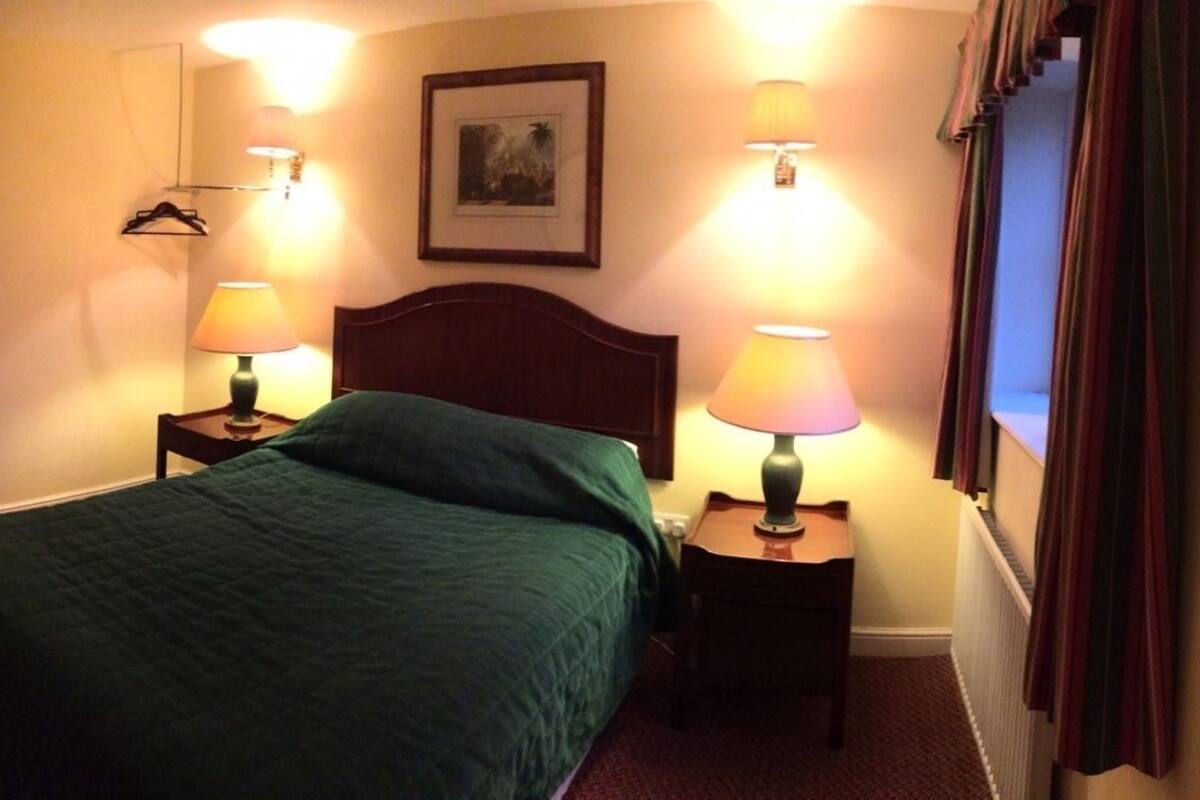 Quarto de hotel com cama, luminárias laterais, espelho, porta e uma pequena mesa com uma xícara e uma caixa de lenços.