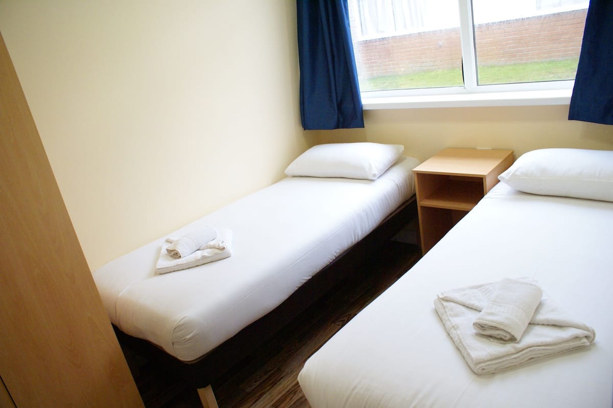 Une chambre avec deux lits simples, chacun équipé d’un oreiller et d’une serviette pliée. Une table de chevet en bois se trouve entre les deux lits.