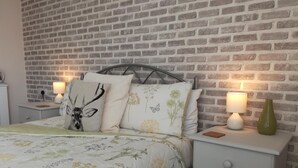 A bedroom with a brick-patterned wall, a bed with floral bedding, two bedside tables with lamps, and a nightstand with a vase. - Saffron House Hotel (Ilfracombe)