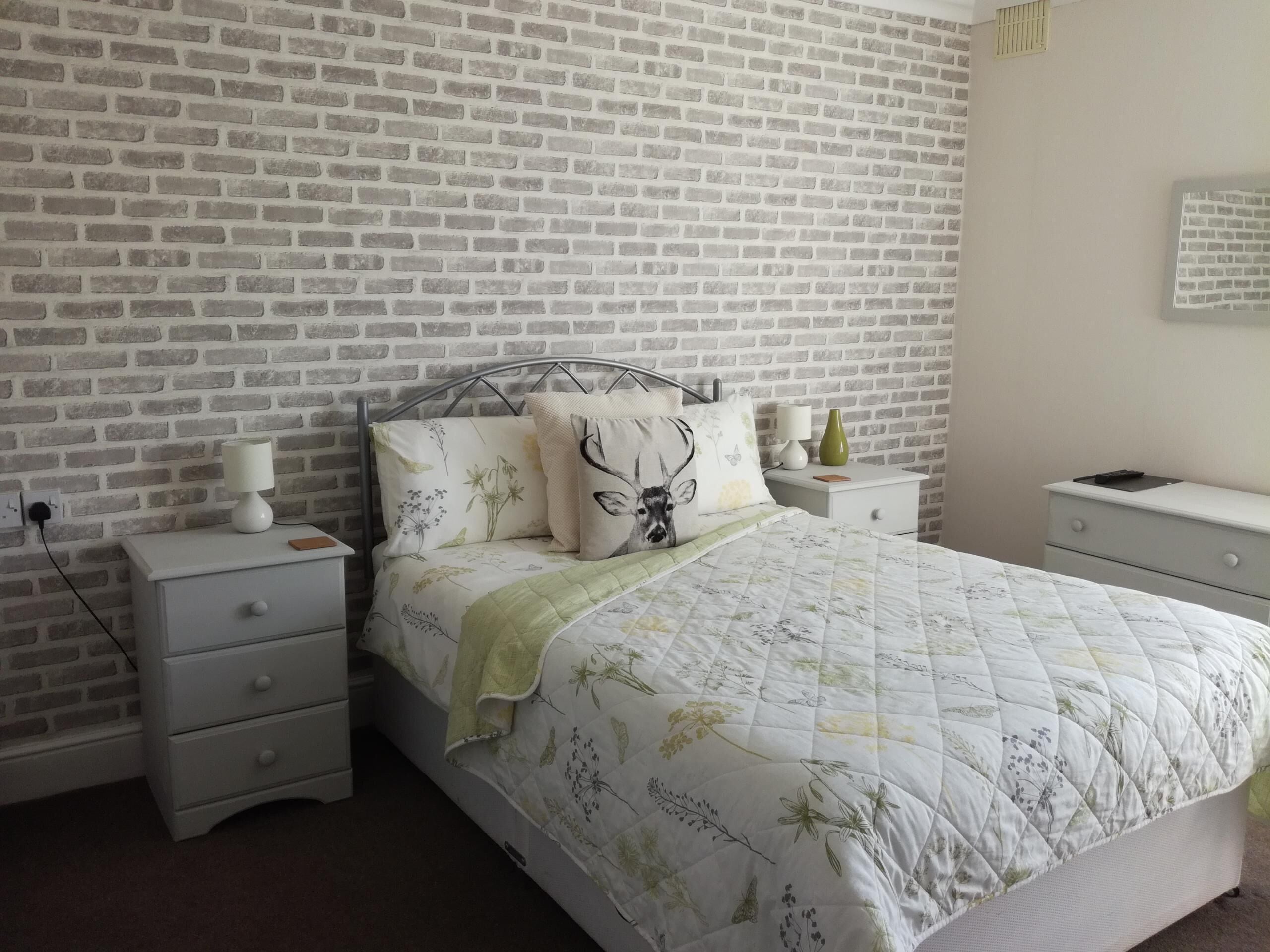 Ein Schlafzimmer mit einer Wand im Ziegelstein-Muster, einem Metallbett, einem Nachttisch mit Lampe und einem Beistelltisch mit Lampe und einer grünen Vase.