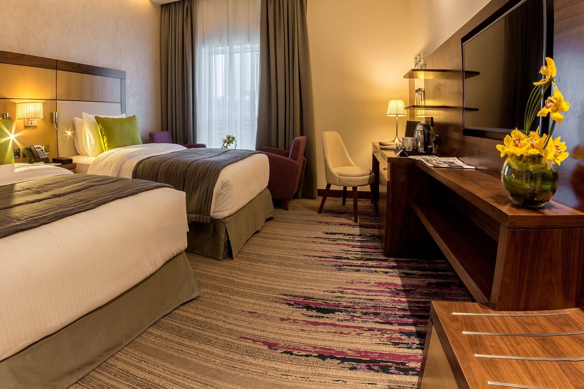 A hotel room with two beds, a desk, a chair, a TV, and a vase with flowers.