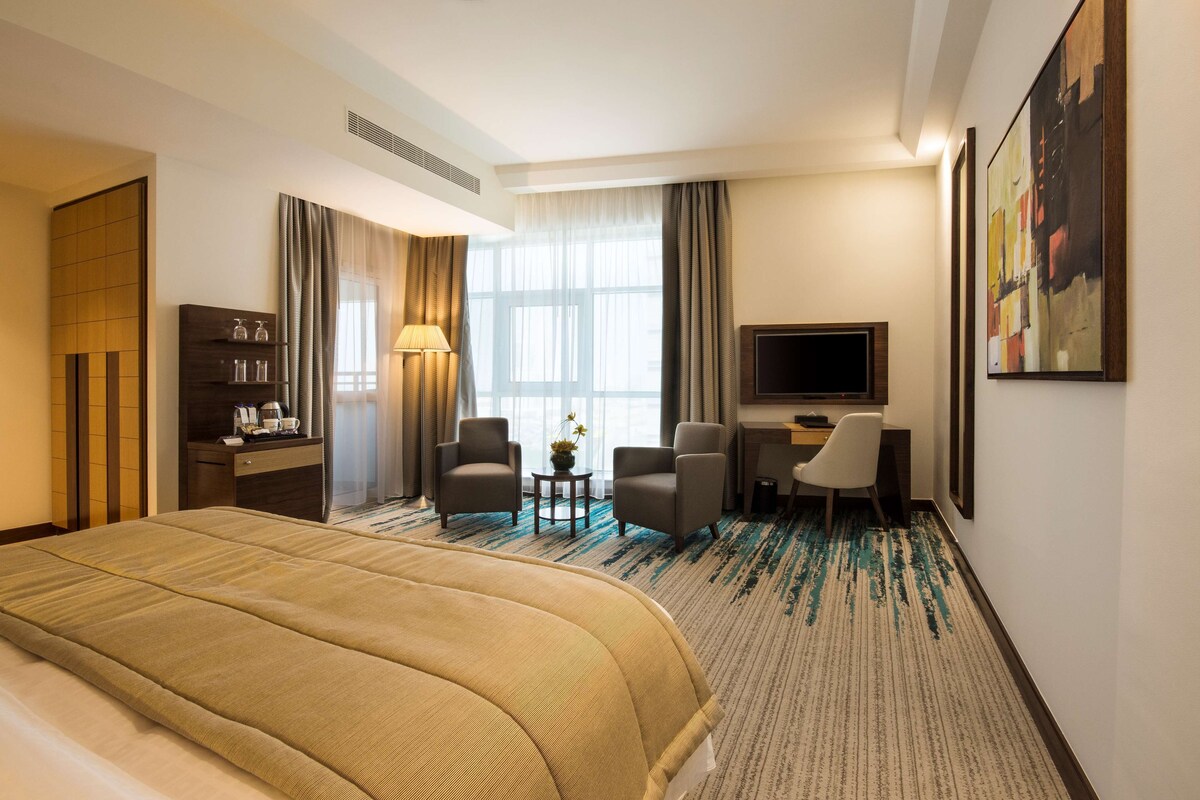 A hotel room with a large bed, two armchairs, a desk with a TV, and a window with curtains.