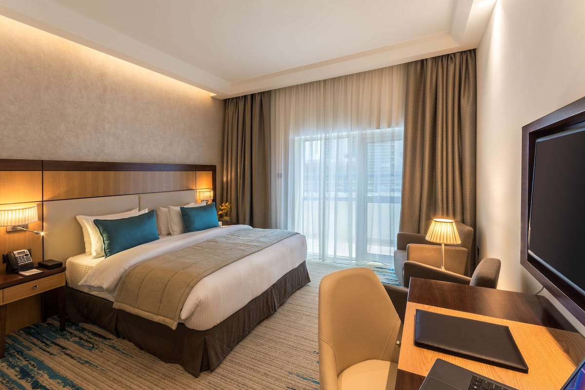 A hotel room with a large bed, a desk with a laptop, a TV, and a window with curtains.