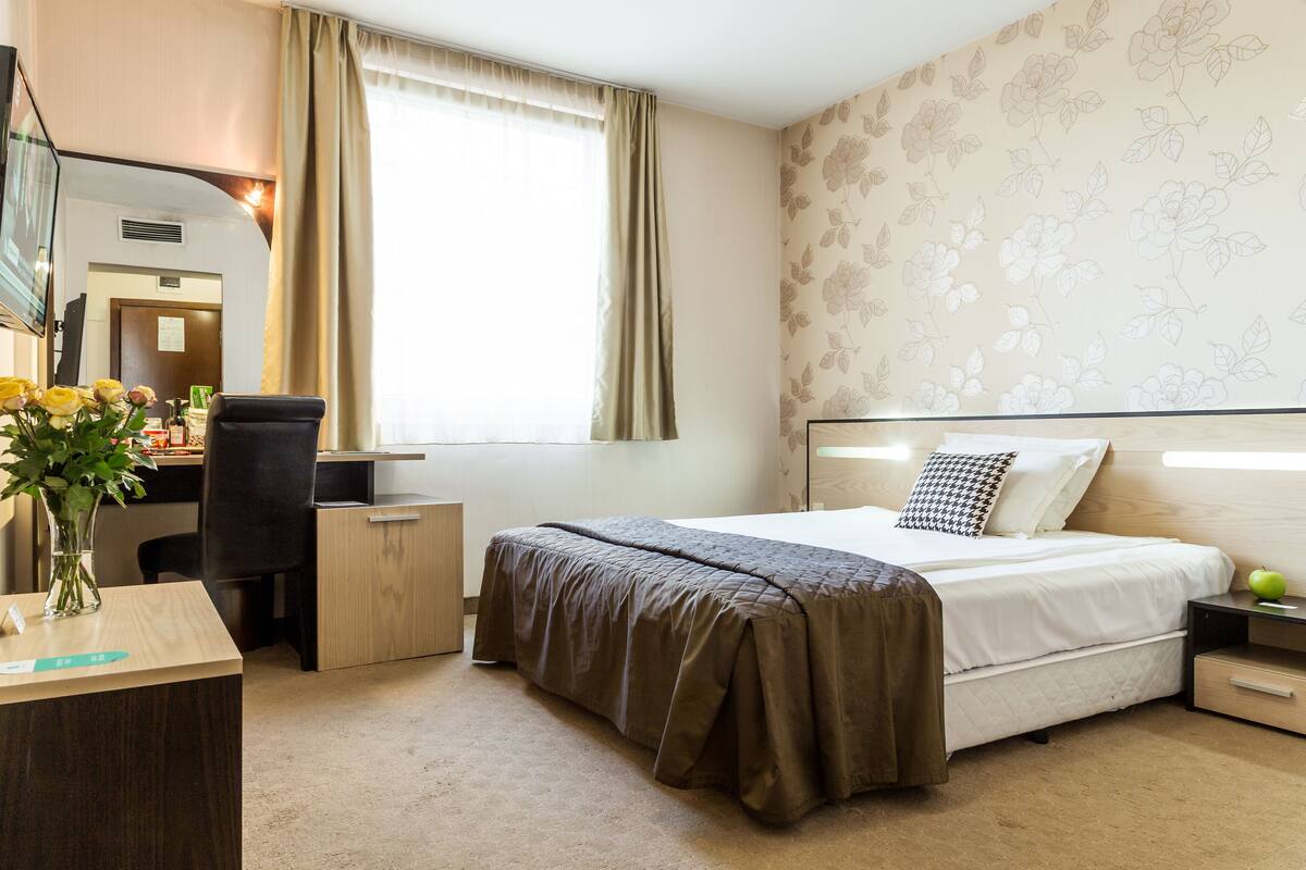 Habitación de hotel con cama, escritorio, silla, mesita, un florero con flores y ventana con cortinas.