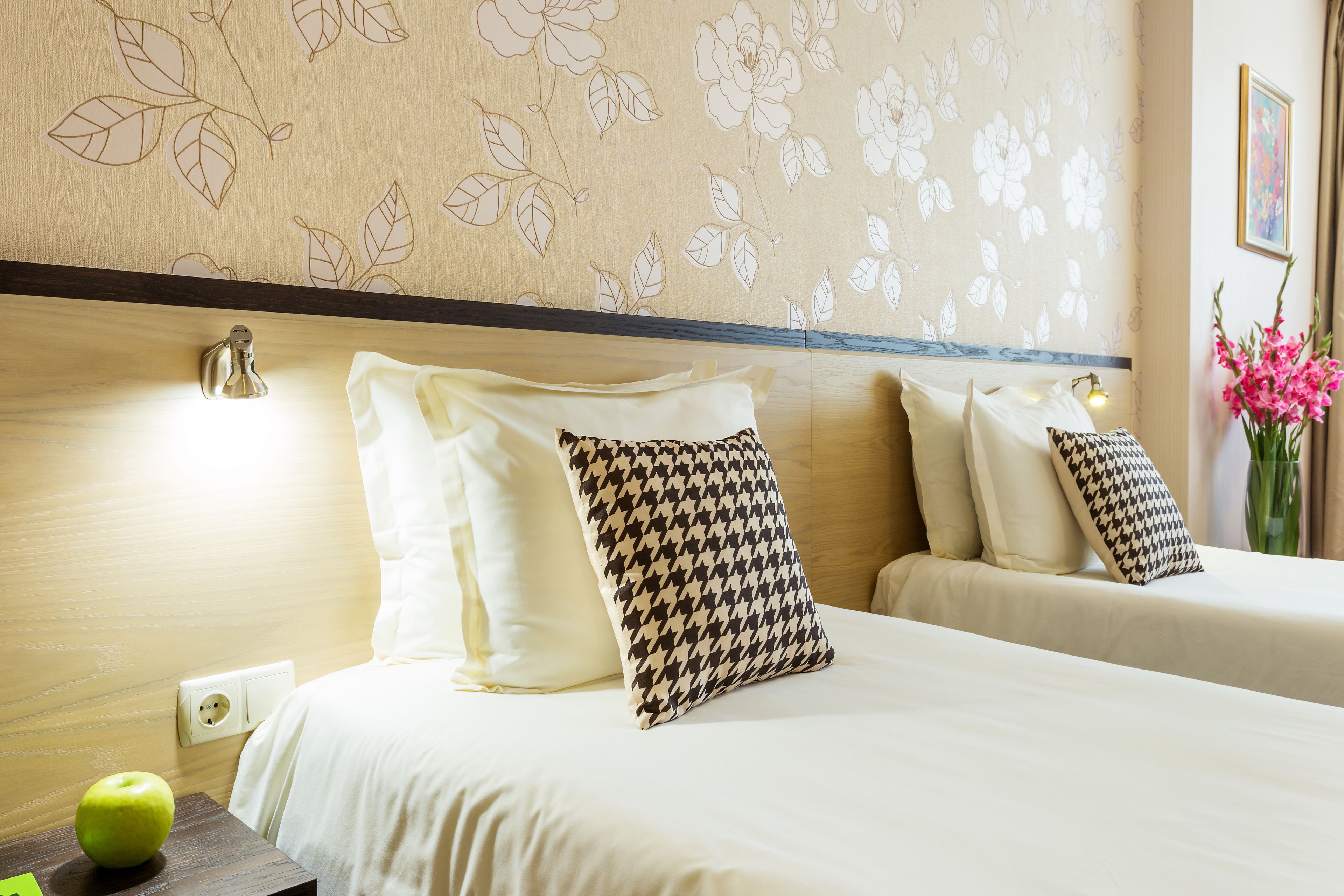 Habitación de hotel con dos camas, cabecero de madera, pared con papel tapiz floral y un florero con flores.