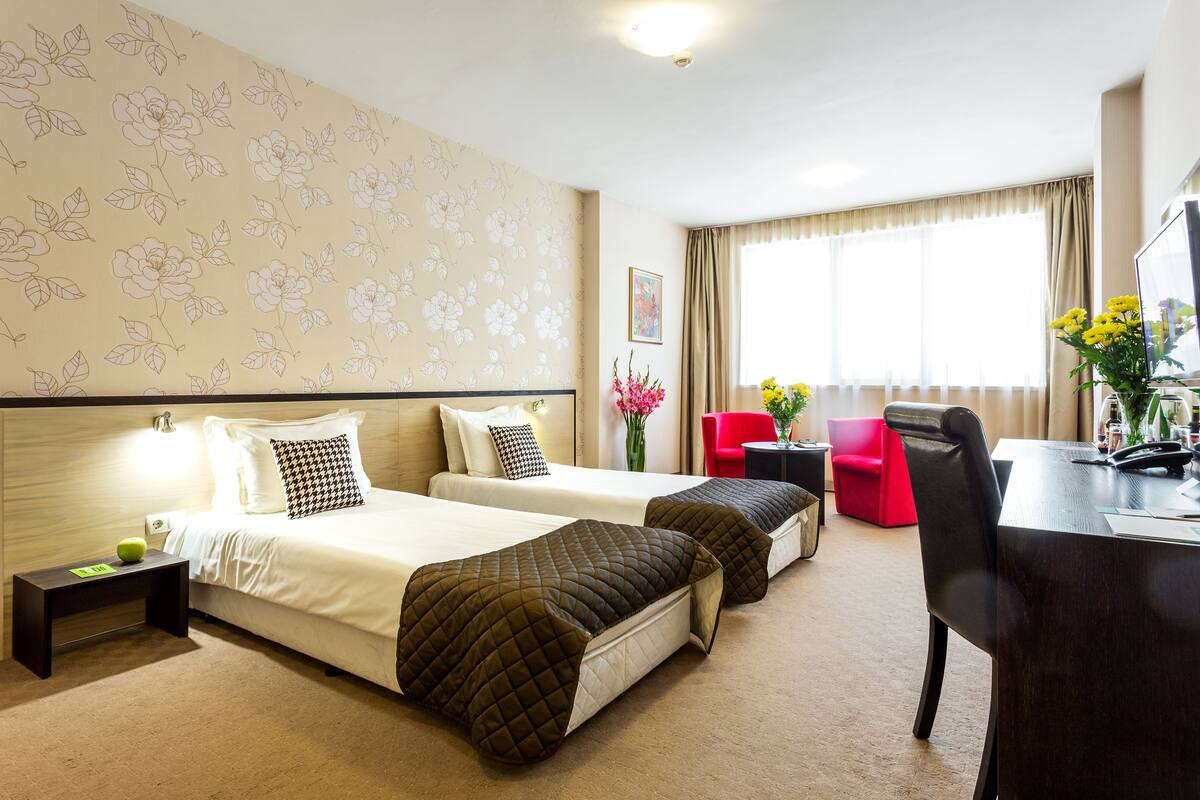 Habitación de hotel con dos camas, un escritorio, una silla, una mesita y un ventanal con cortinas.