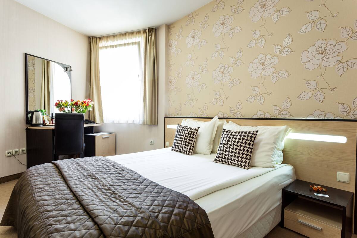 Una habitación de hotel con cama, escritorio, silla, mesita de noche y papel tapiz floral.