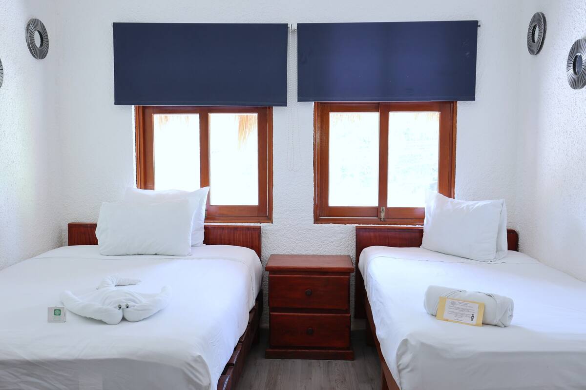 Habitació doble amb dos llits, una nitrera de fusta i una finestra amb una persiana blau fosc.