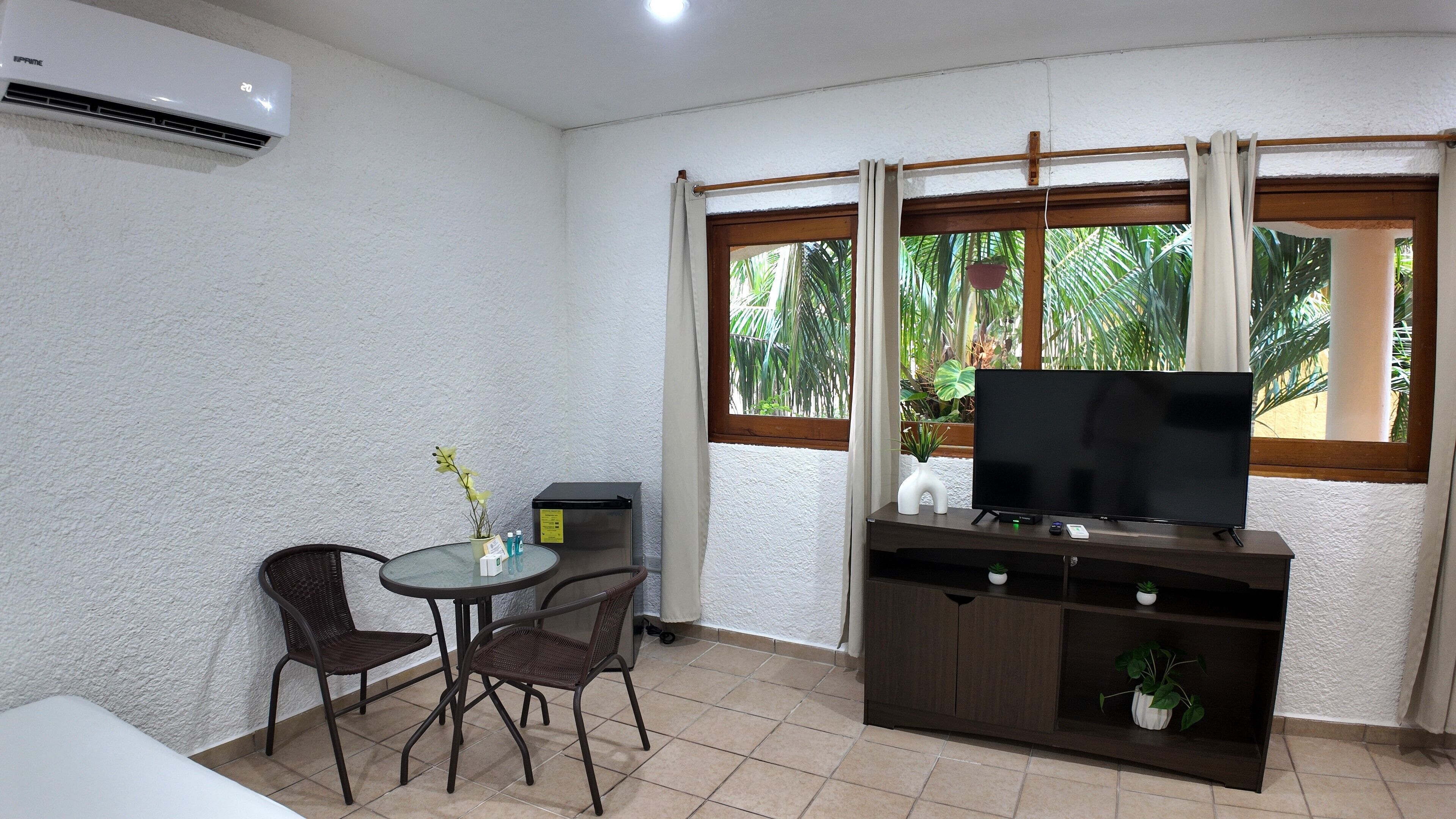 Um quarto pequeno com televisão, uma mesa com cadeiras, um armário e uma janela com cortinas.