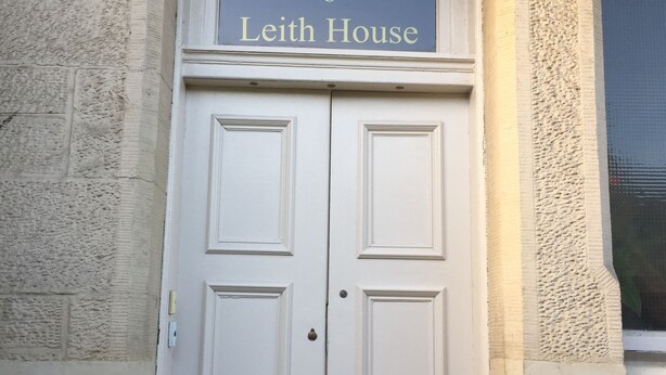 Una puerta doble blanca con pomo de latón y una pequeña ventana encima. La puerta tiene la leyenda 'Leith House' con el número '6' arriba.