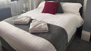 Cama bem arrumada com edredom branco e cinza, duas toalhas dobradas e um travesseiro vermelho.