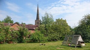 A church with a tall spire, surrounded by greenery and a wooden playhouse. - Church Farm (Solihull)