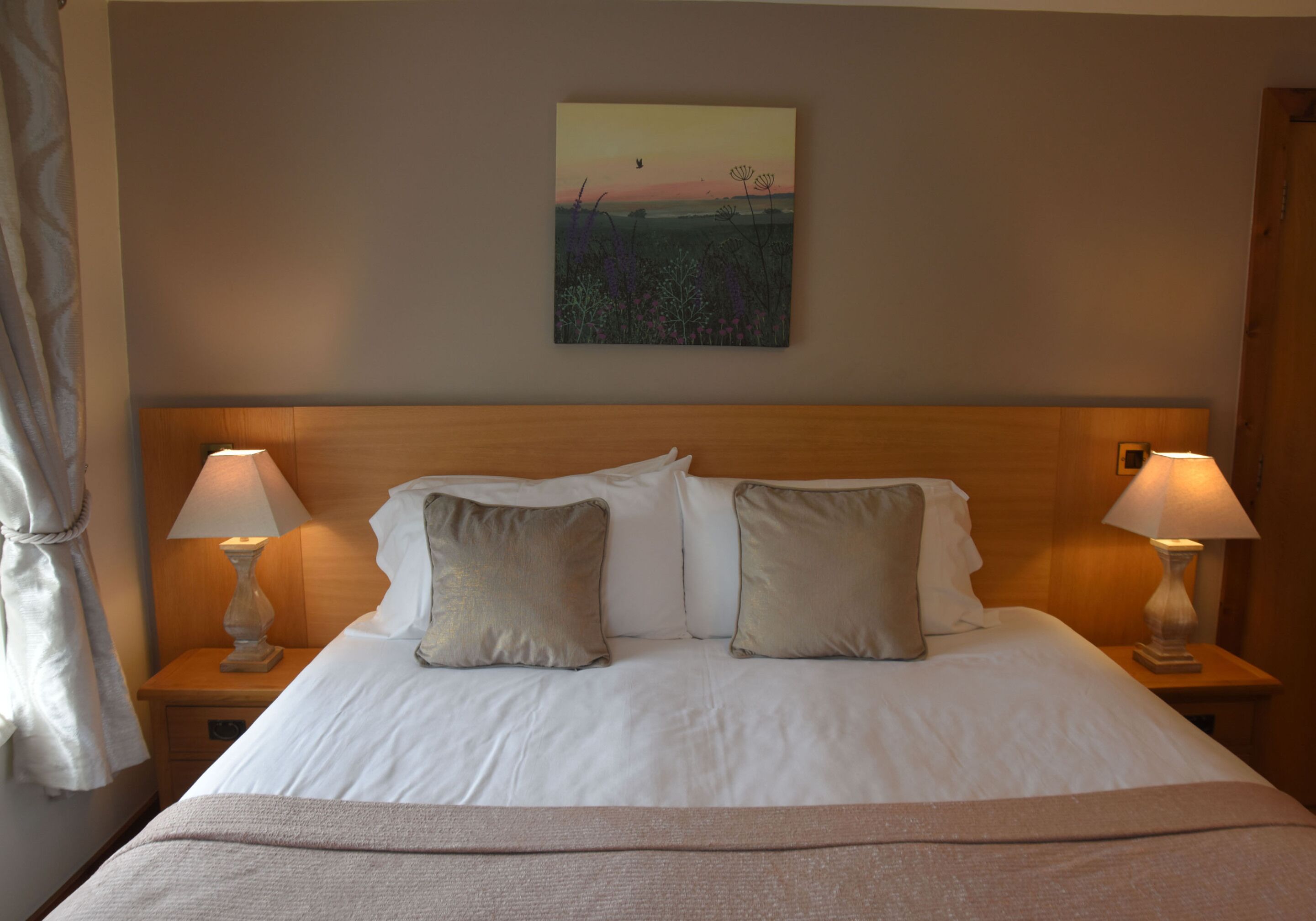 A neatly made bed with two pillows, two bedside lamps, and a painting of a sunset on the wall.
