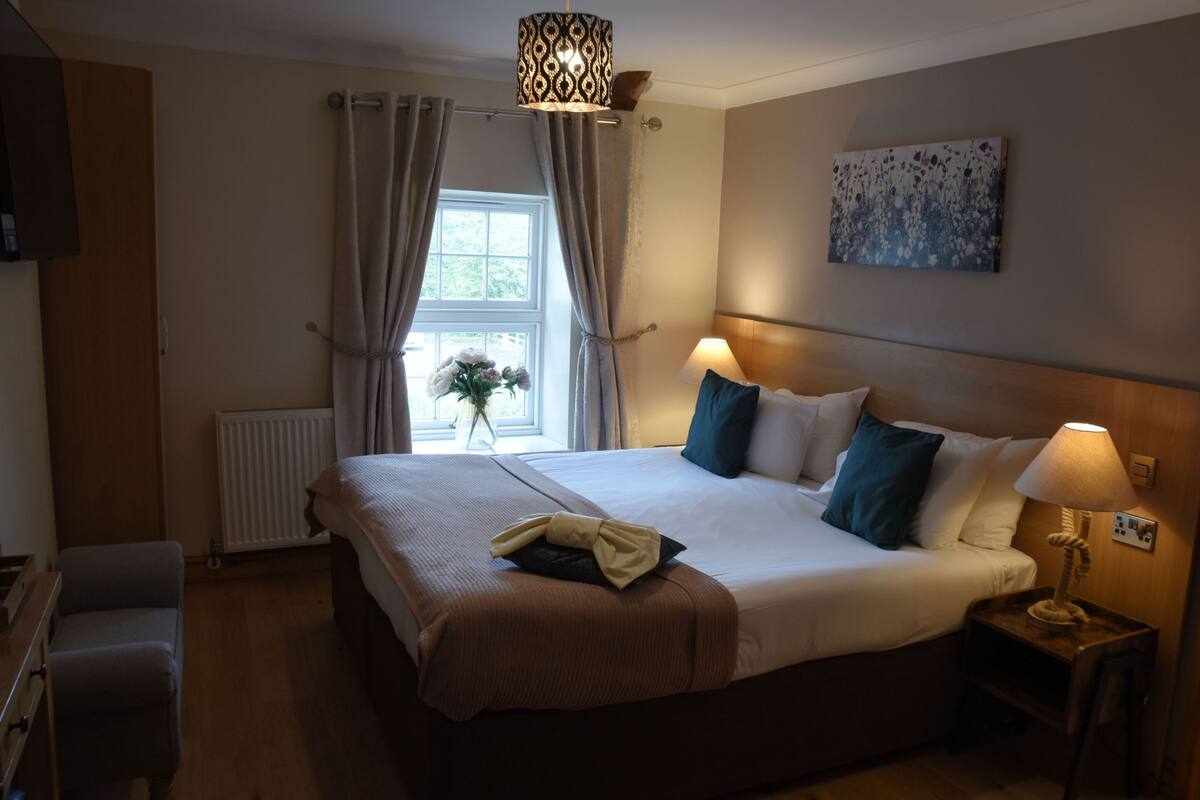 A neatly made bed with a brown bedspread, a bedside lamp, and a window with curtains.