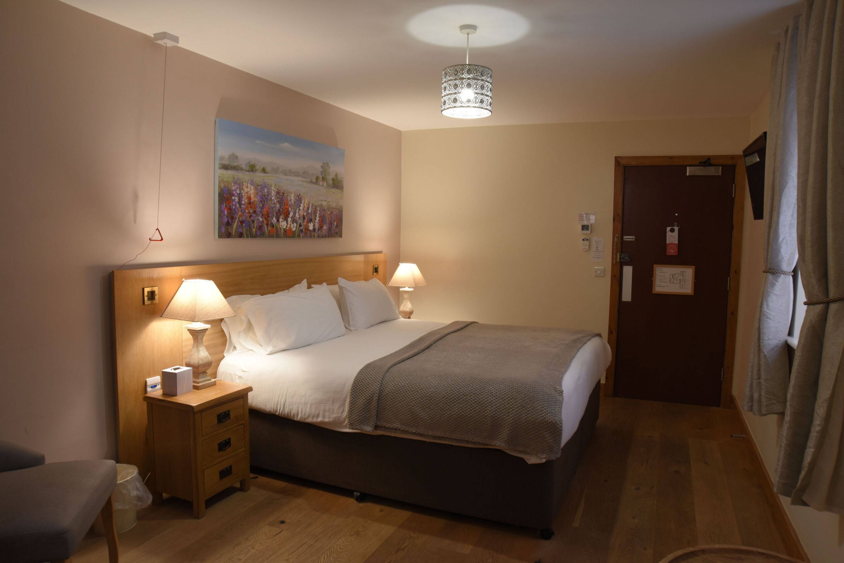 A hotel room with a bed, two bedside lamps, a nightstand, a painting on the wall, and a door with a sign.