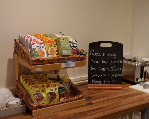 A breakfast buffet with cereal and coffee options, a sign offering tea, coffee, juices, toast, cereal, and yoghurts, and a toaster on the counter.