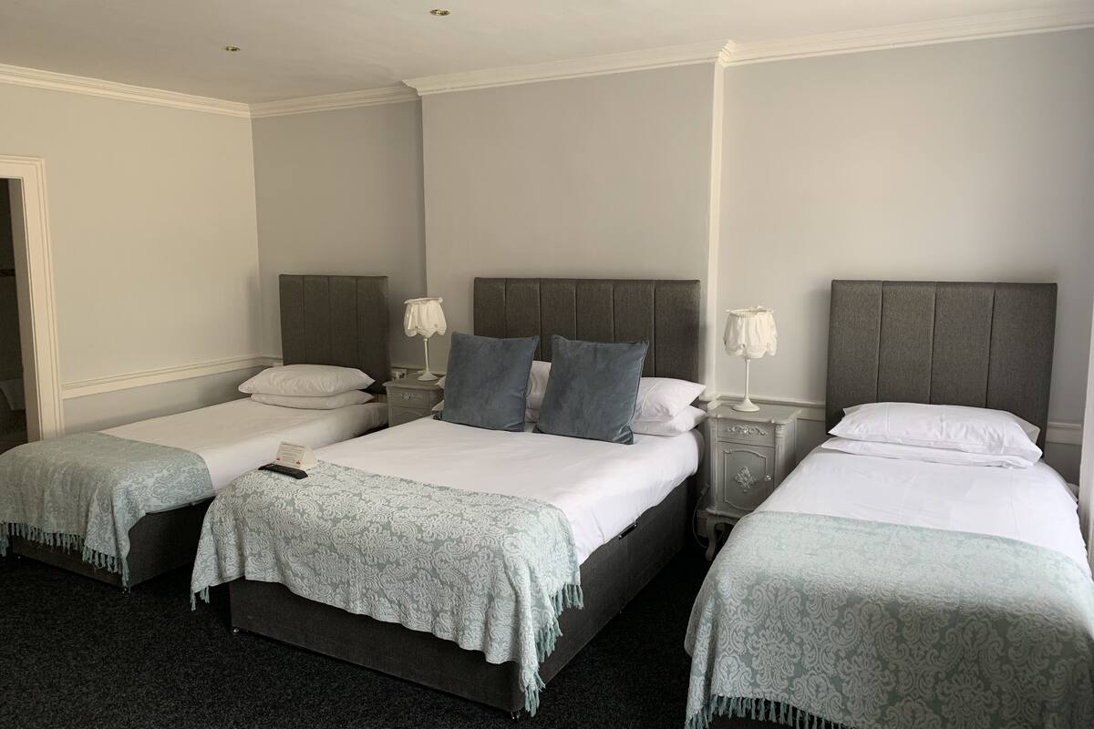 A hotel room with three single beds, each with a grey headboard and white bedding. There are bedside tables with lamps and a small drawer. The room has a carpeted floor and white walls.