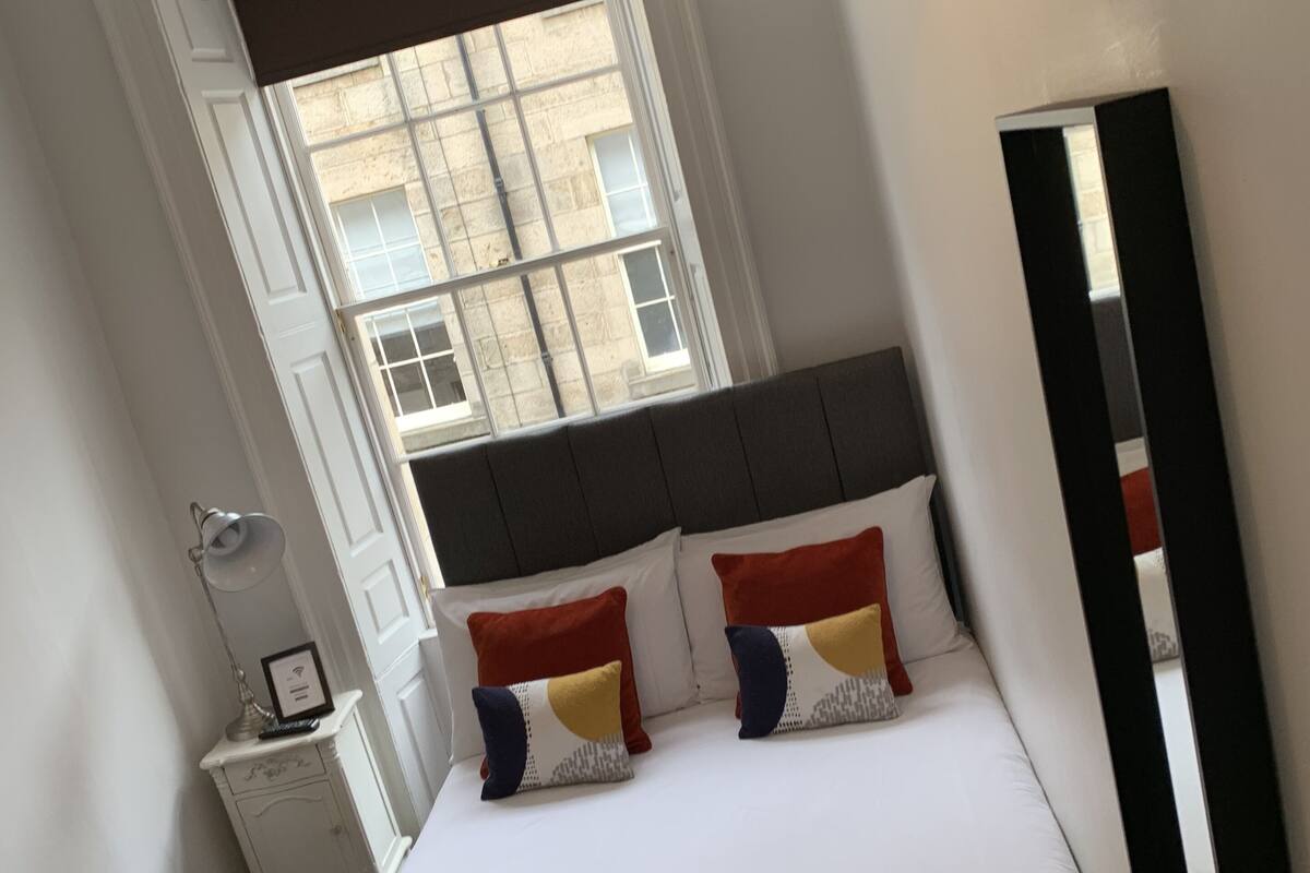 A hotel room with a bed, a bedside table, a mirror, and a window with a view of buildings.