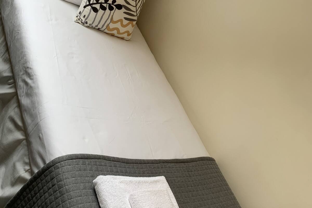 A neatly made bed with a white duvet, a gray quilted blanket, and a white towel folded on the bed.