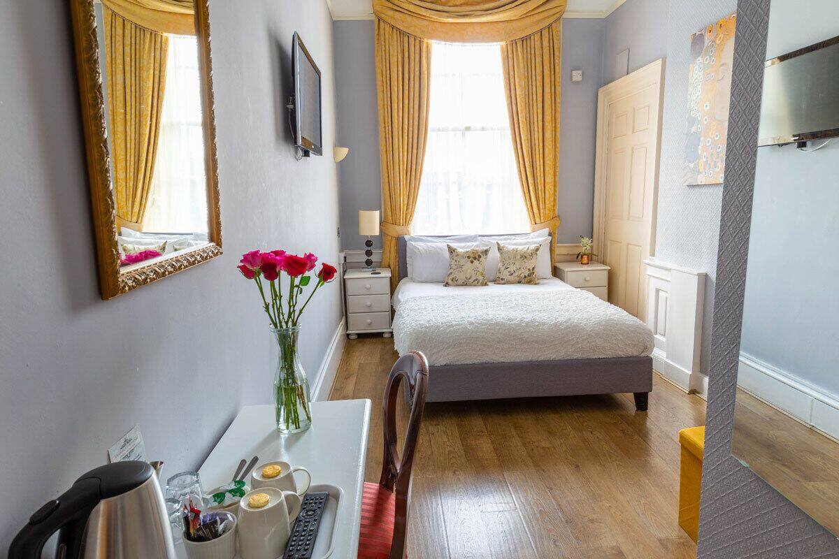 Quarto com cama, mesas de cabeceira, escrivaninha com cadeira, espelho e janela com cortinas.