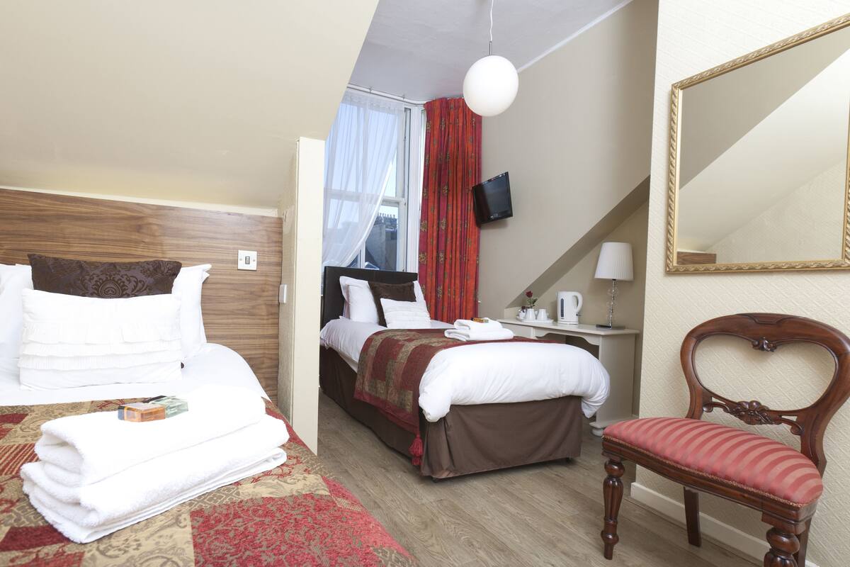 Quarto de hotel com cama, poltrona, escrivaninha, televisão e espelho.