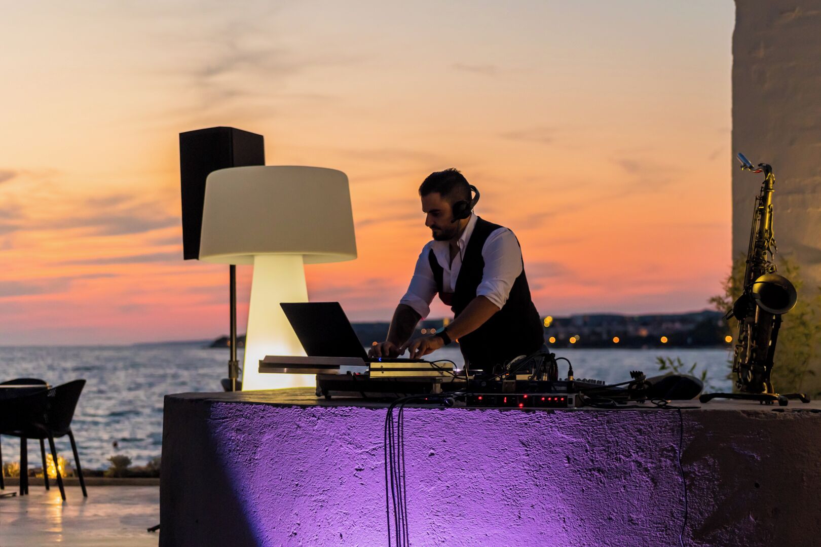A DJ setup with a laptop, turntables, and a saxophone at a seaside venue with a sunset backdrop.