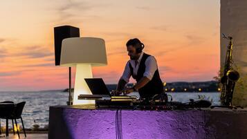 A DJ setup with a laptop, turntables, and a saxophone at a seaside venue with a sunset backdrop.