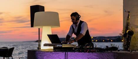 A DJ setup with a laptop, turntables, and a saxophone at a seaside venue with a sunset backdrop.