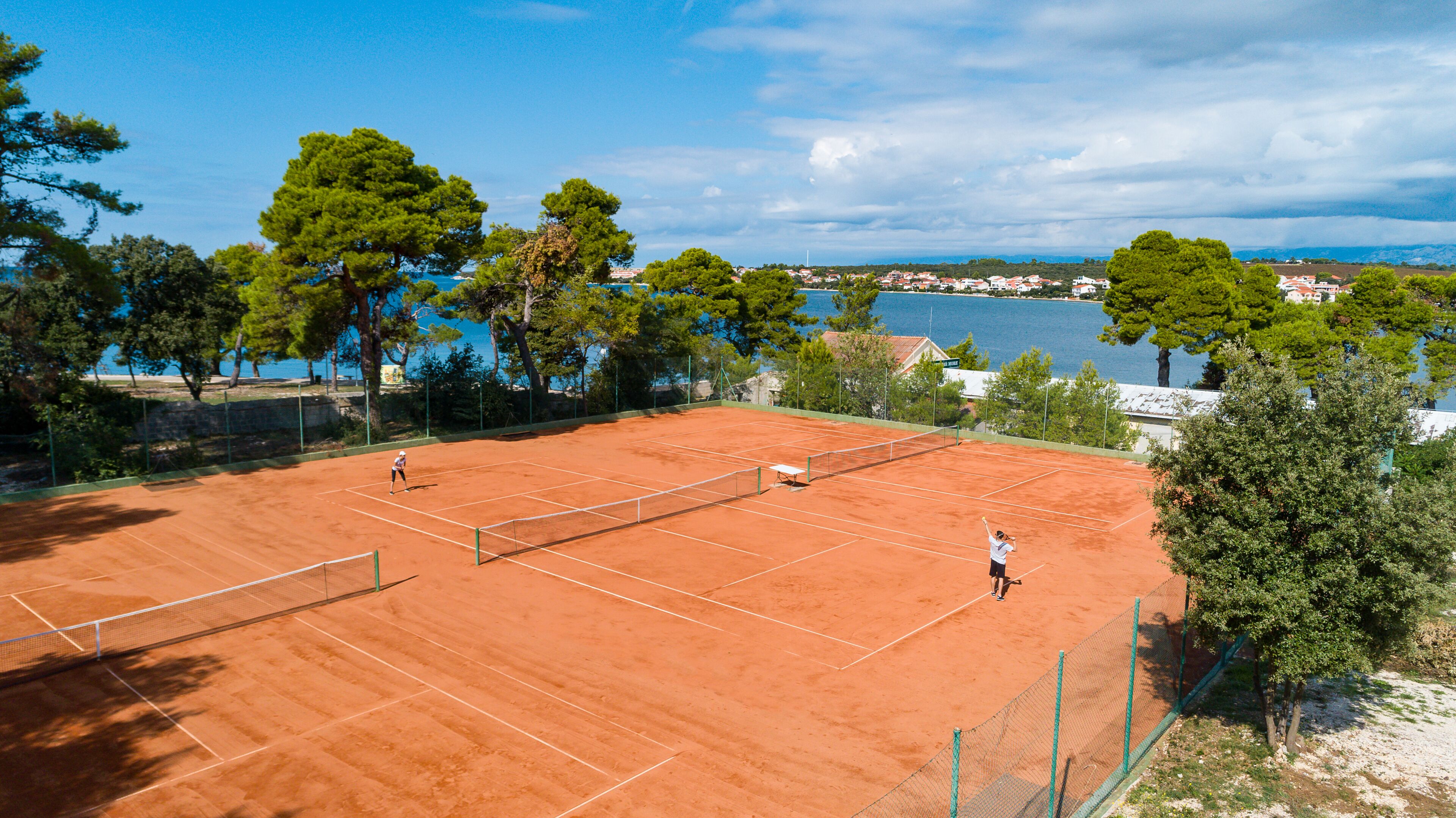 A tennis court with a net, surrounded by trees and a view of a body of water and buildings in the distance.