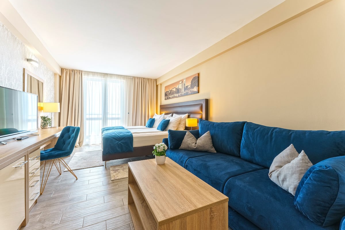 A modern hotel room with a wooden desk, a blue sofa, a bed with matching pillows, and a cityscape painting on the wall.