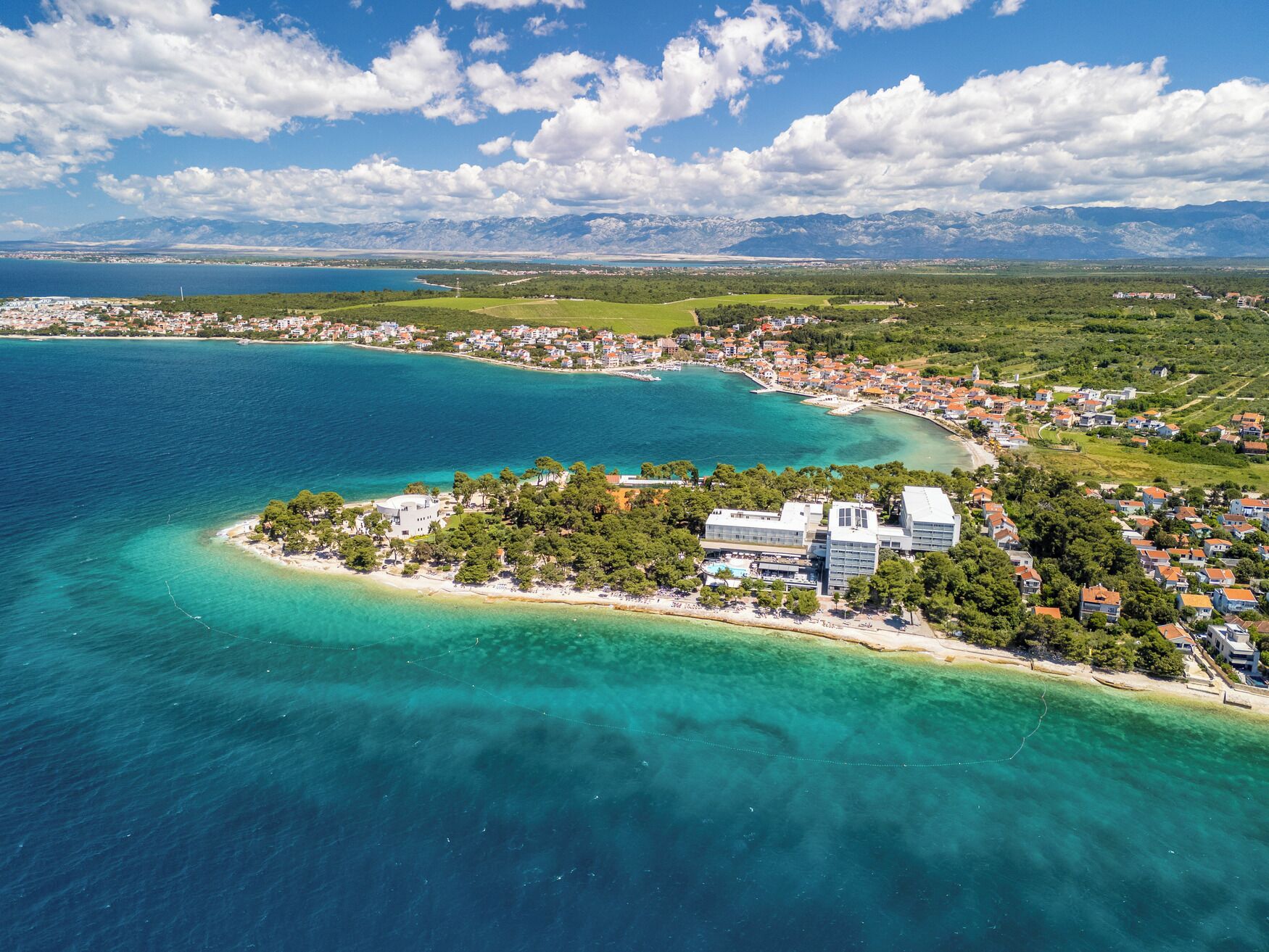 An aerial view of a coastal town with a prominent building surrounded by clear blue waters and a mountainous backdrop.