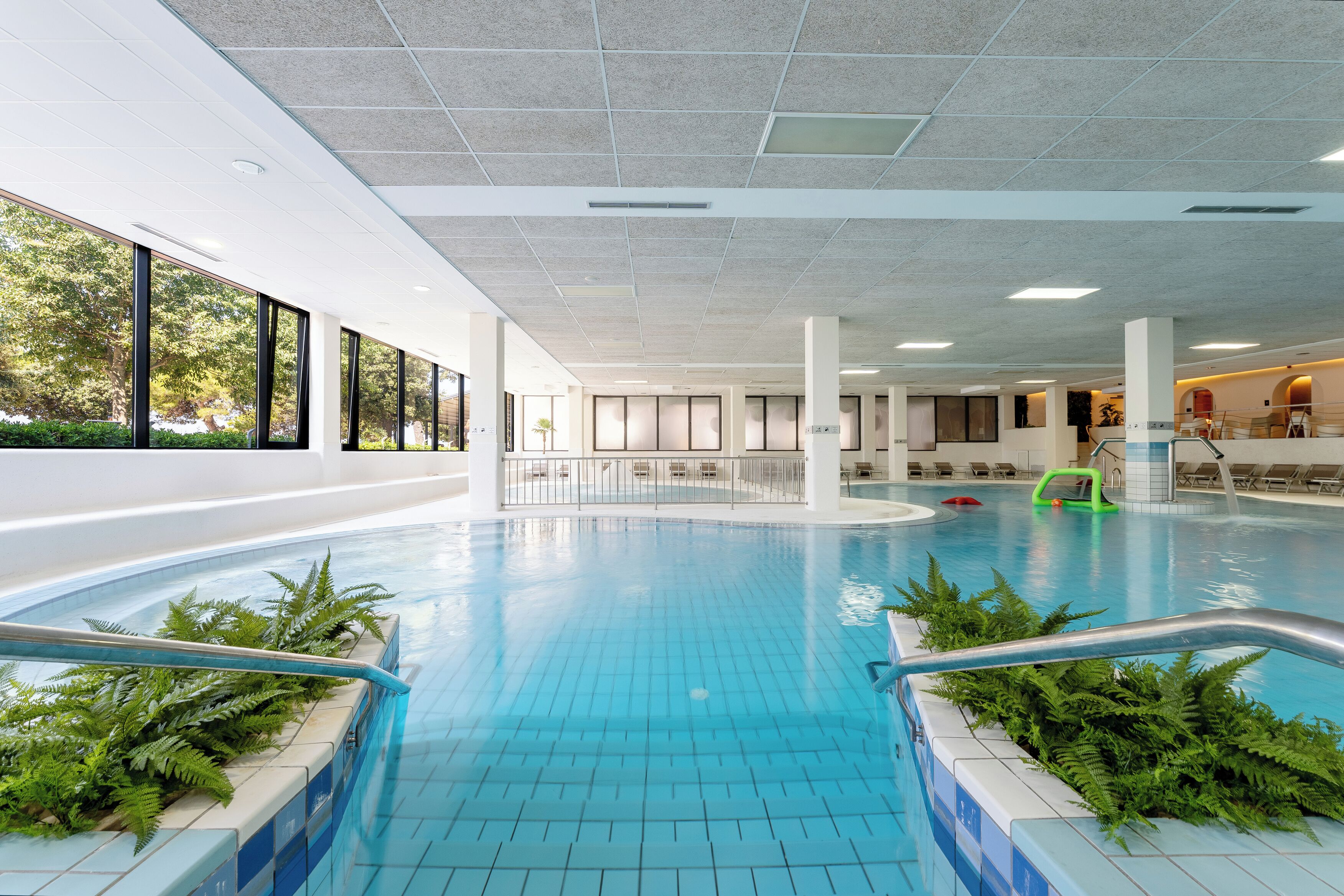 An indoor swimming pool with clear blue water, surrounded by white tiled walls and large windows.