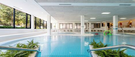 An indoor swimming pool with clear blue water, surrounded by white tiled walls and large windows.
