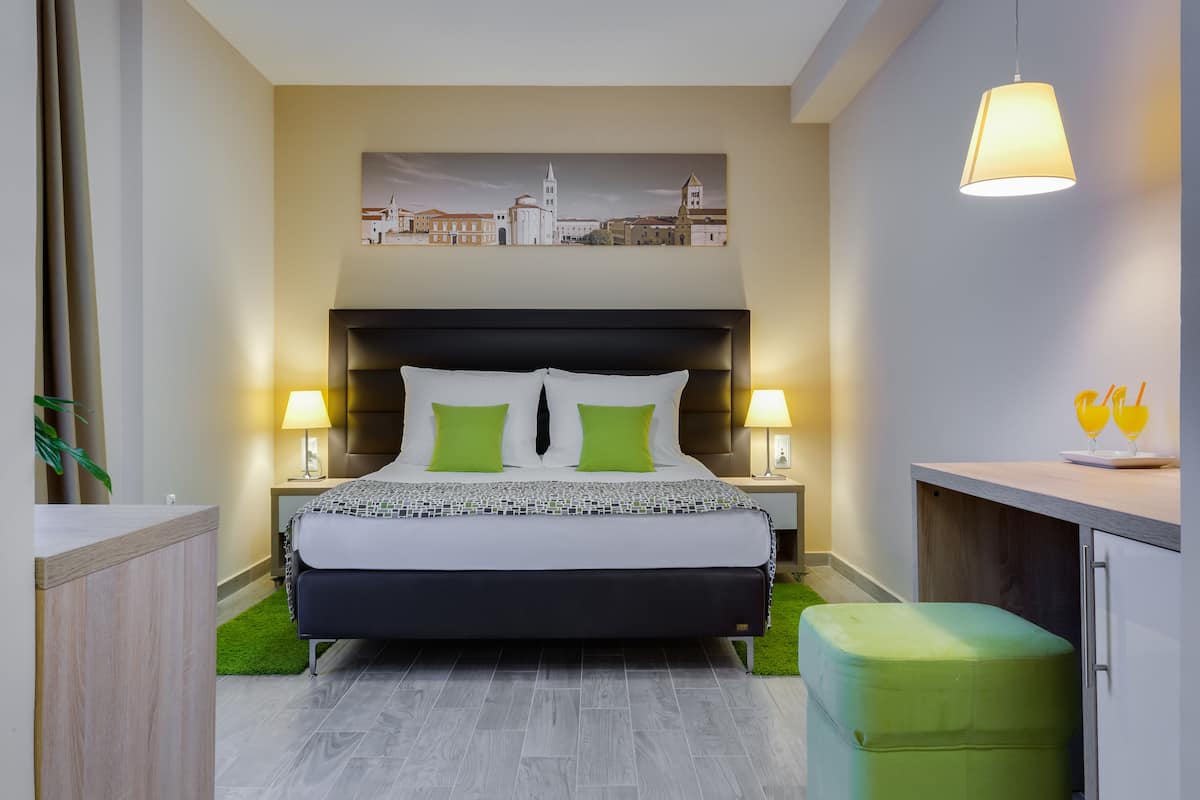 A modern hotel room with a large bed, two bedside lamps, a green ottoman, and a cityscape painting on the wall.