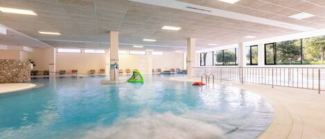 An indoor swimming pool with clear water, a stone wall, and a view of greenery outside.