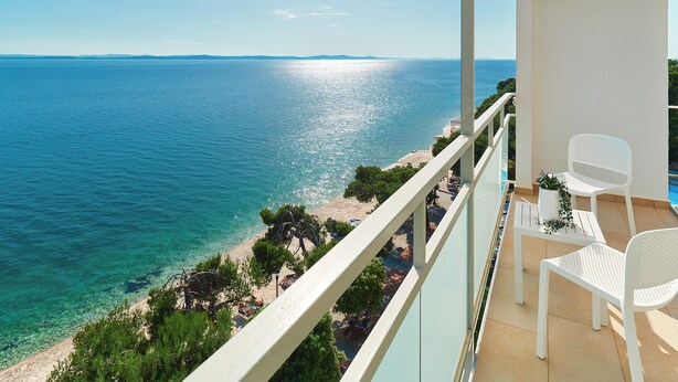 Un balcon avec des chaises blanches et une petite table, donnant sur une plage et la mer.
