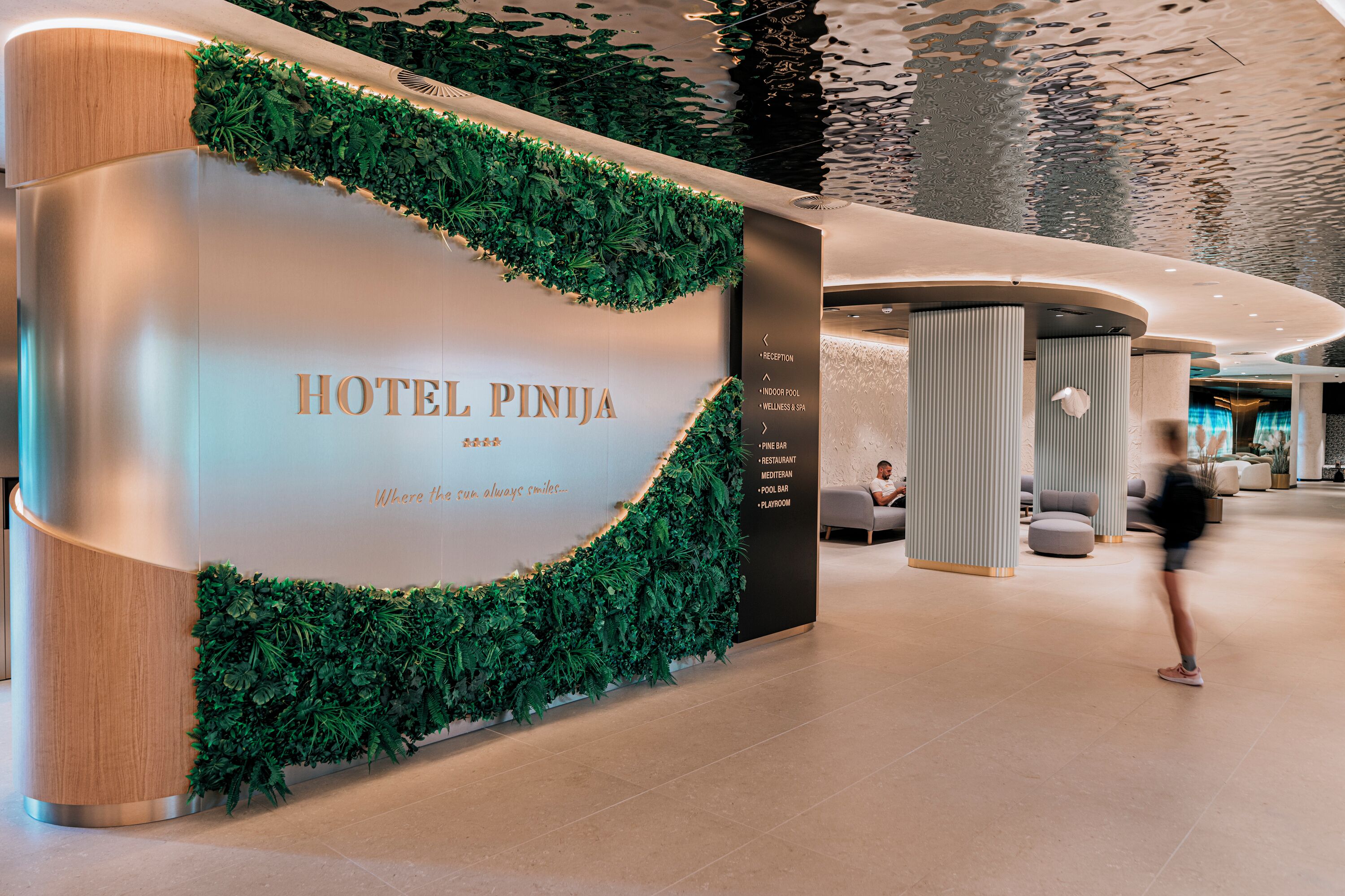 A hotel reception area with a modern design, including a reception desk, a wall with greenery, and a list of amenities such as a pool, restaurant, and wellness center.