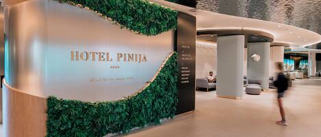 A hotel reception area with a modern design, including a reception desk, a wall with greenery, and a list of amenities such as a pool, restaurant, and wellness center.