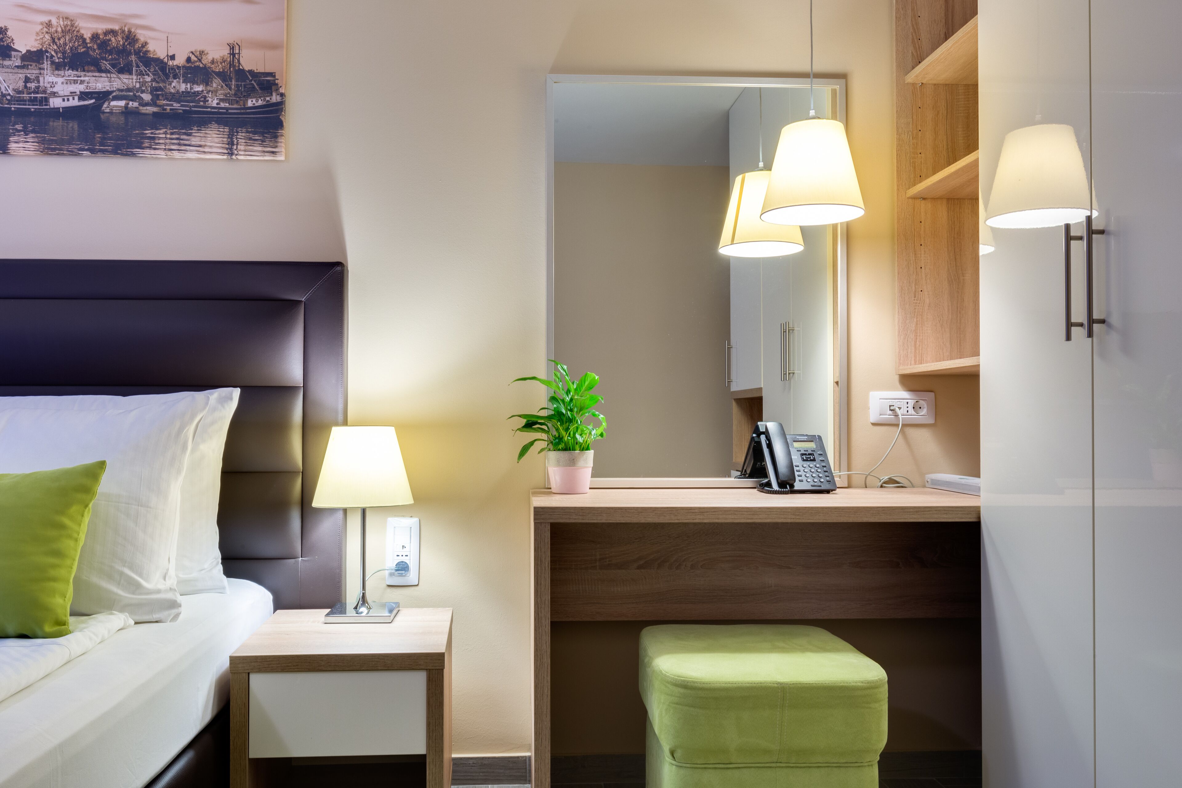 Et hotelværelse med en seng, et sengebord, en lampe, en lille grøn plante, et skrivebord med en telefon og et spejl.