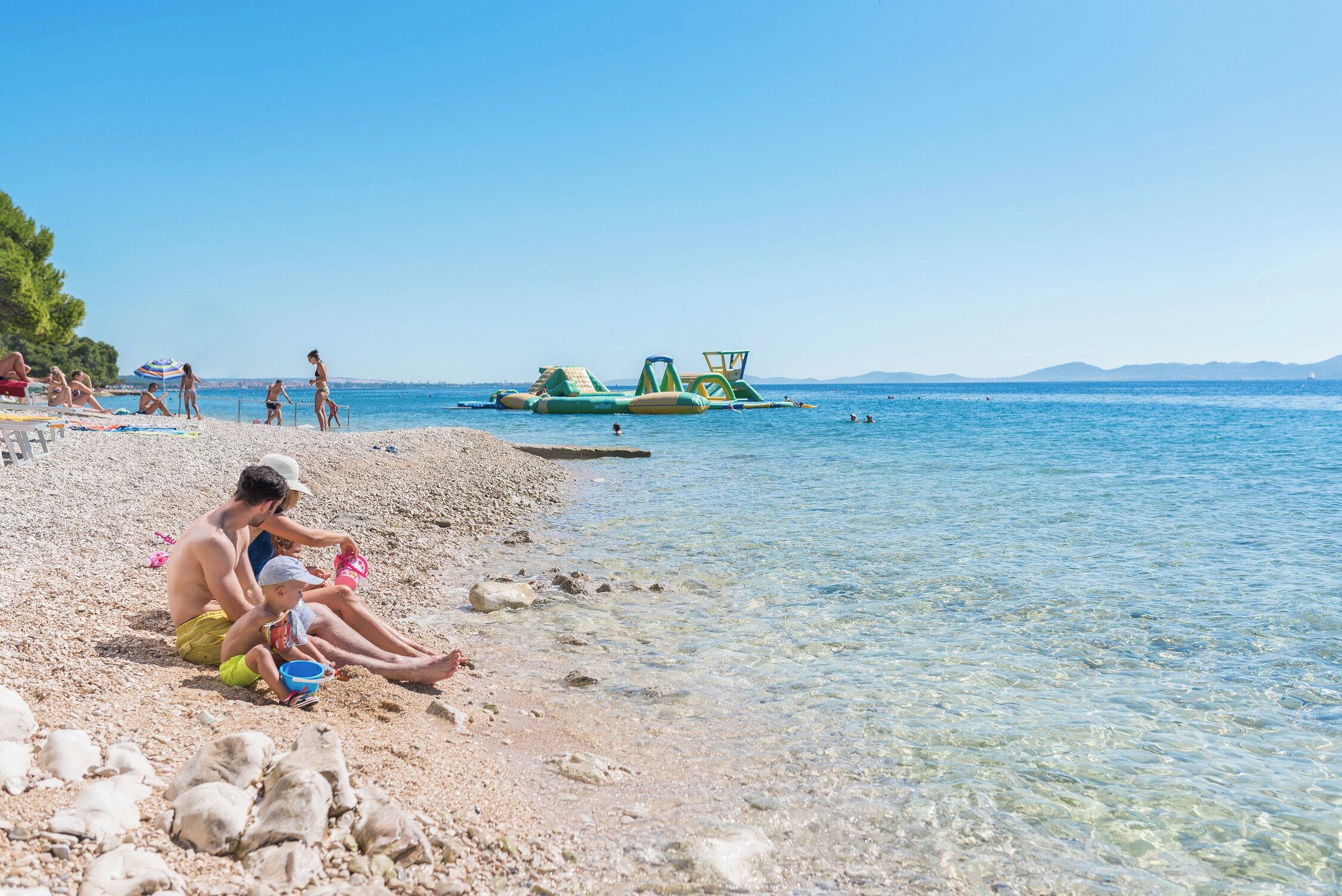 A beach with clear water, people relaxing, and inflatable water structures.