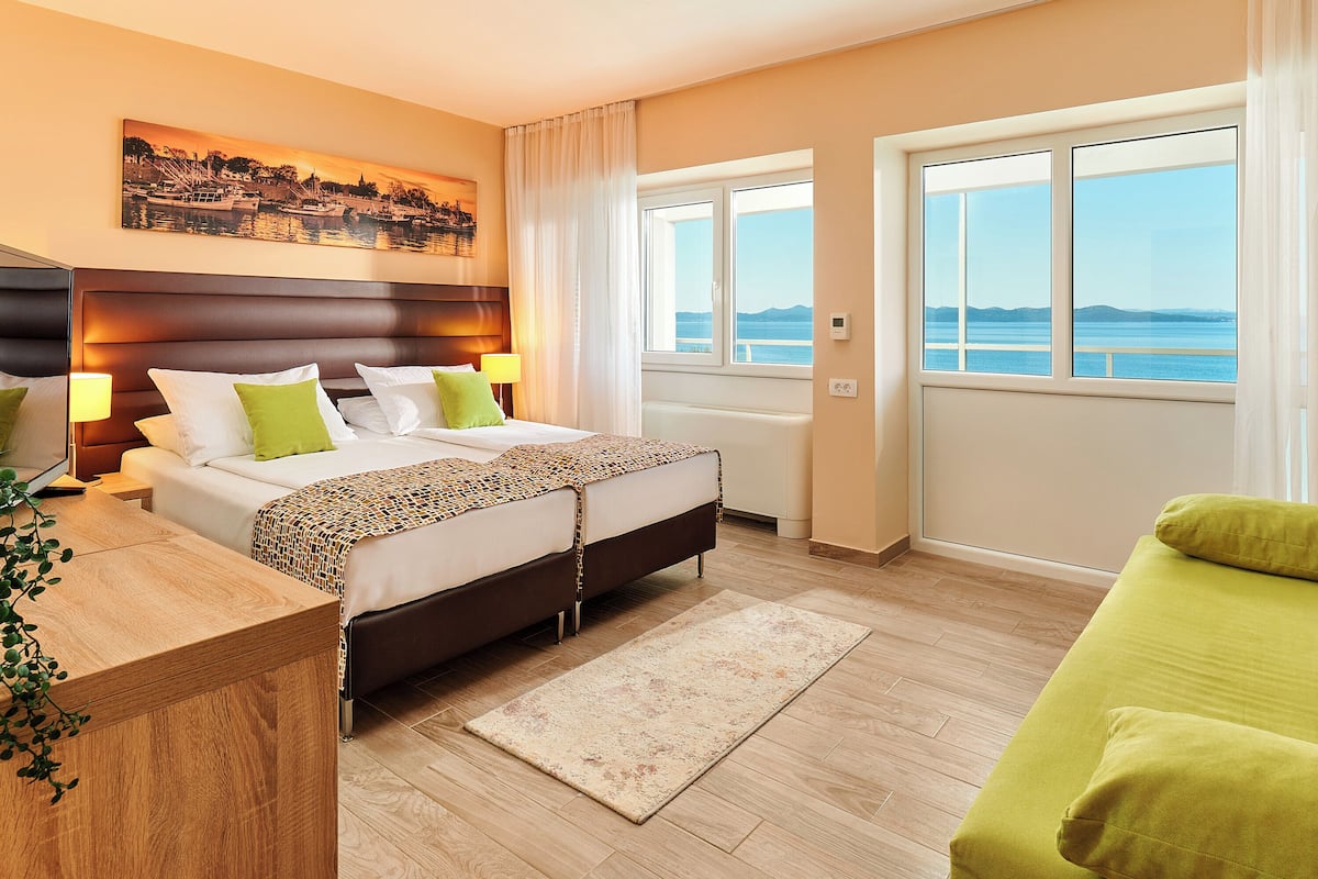 A hotel room with a bed, a desk, a window with a view of the sea, and a painting on the wall.