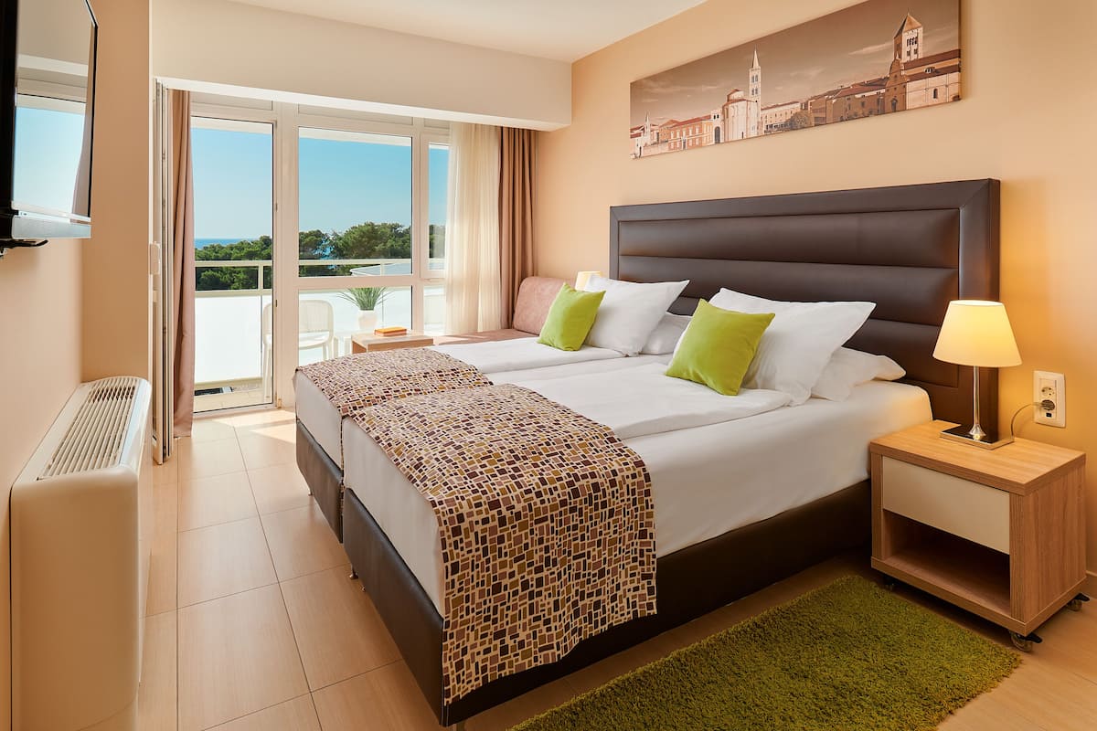A hotel room with a large bed, a bedside table with a lamp, a radiator, and a view of the outdoors through a sliding glass door.