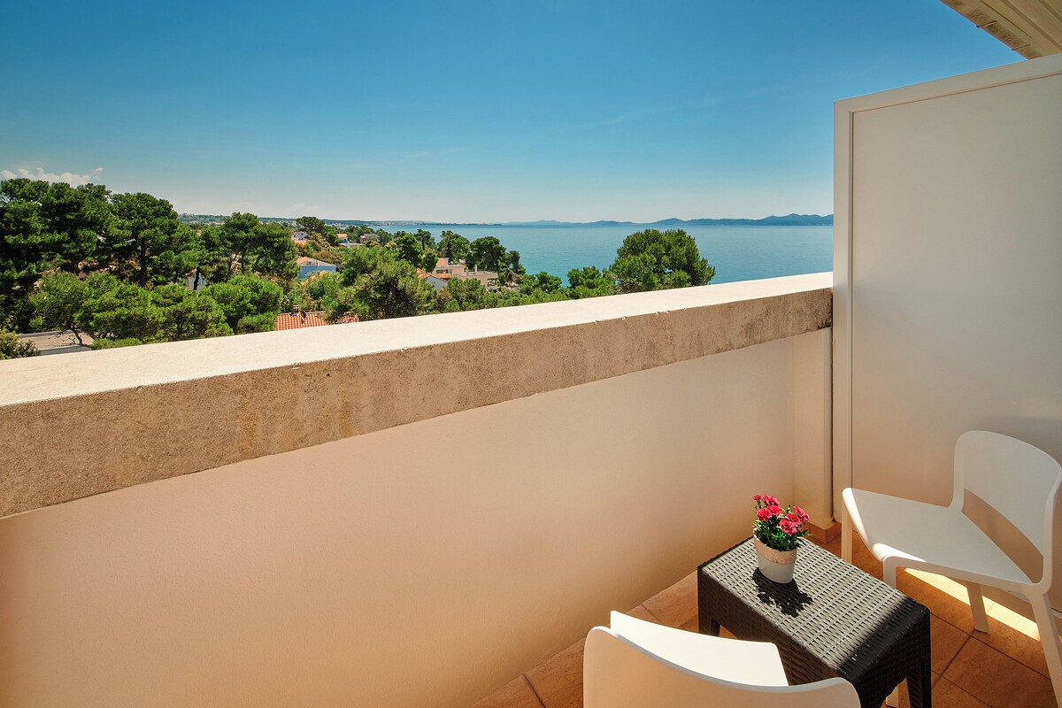 A balcony with a view of the sea, a table with a potted plant, and white chairs.