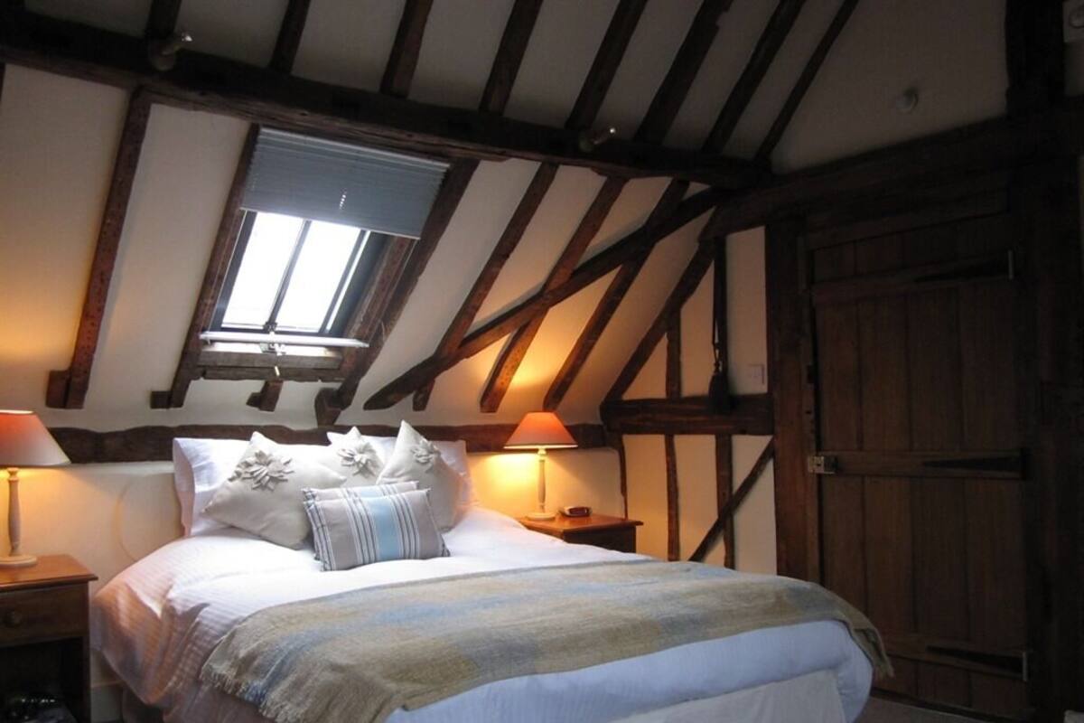 A bedroom with a sloped ceiling, wooden beams, a large window, a bed with white bedding and decorative pillows, two bedside lamps, and a wooden wardrobe.